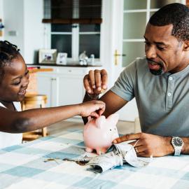 Un hombre y una niña sentados en una mesa con una alcancía, ahorrando juntos para su futuro.