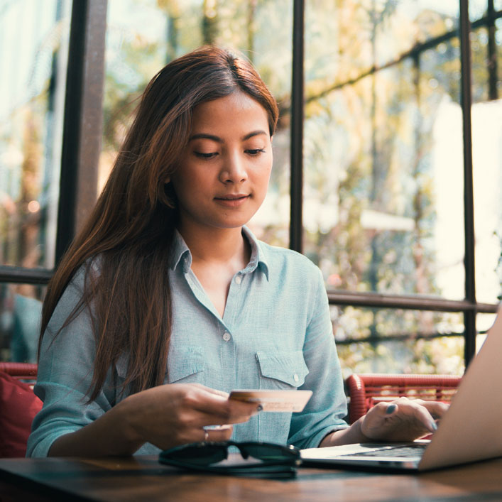 Una mujer sentada en una mesa, usando una computadora portátil y sosteniendo una tarjeta de crédito para transacciones en línea.